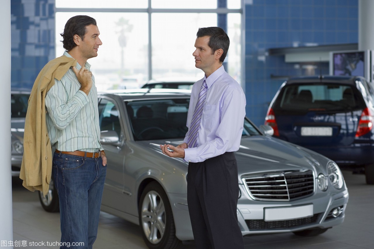 汽车销售员与男顾客在展厅里的新银轿车旁交谈,微笑,侧视